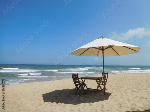 A secluded sandy beach scene with white breakers in background, foreground of wooden table and chairs with large cream parasol. © Julie