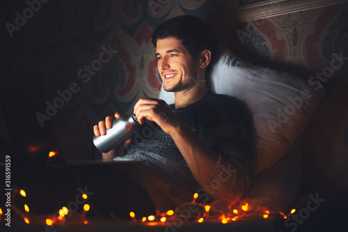 Night portrait of young smiling man lying on pillow on bed in dark room at home, holding reusable aluminum thermo water bottle, looking in laptop.