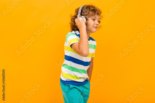 Little boy with curly hair in colourful t-shirt and shorts listen to music with big earphones isolated on yellow background