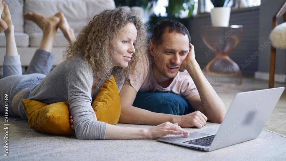 Couple watching computer in pajamas. Cropped portrait man and woman relaxing on carpet spending weekends morning together using laptop, browsing or online shopping in modern European flat Couple watching computer in pajamas. Cropped portrait man and woman relaxing on carpet spending weekends morning together using laptop, browsing or online shopping in modern European flat