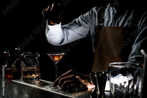 Male bartender spraying on a brown alcoholic drink in a martini glass with orage zest juice