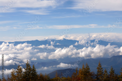 High, blue mountains partly covered in clouds. Some blue sky and a forest in the foreground