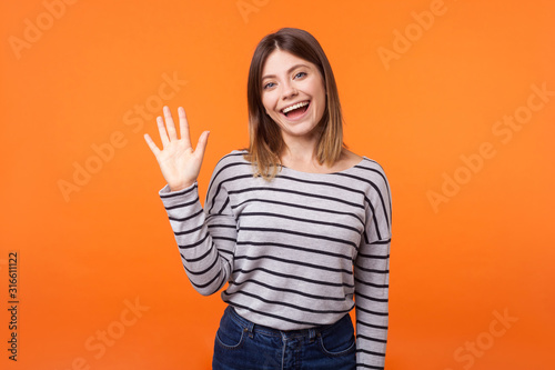 Hello! Portrait of adorable friendly woman with brown hair in long sleeve shirt standing waving hand, looking at camera with engaging toothy smile. indoor studio shot isolated on orange background