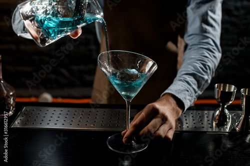 Professional male bartender pouring a blue alcoholic cocktail from the measuring cup with strainer to a martini glass