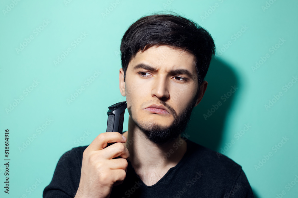 Studio portrait of young man with half shaved face, holding electric shaver trimmer on background of aqua menthe color. Studio portrait of young man with half shaved face, holding electric shaver trimmer on background of aqua menthe color.
