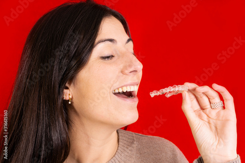 beautiful brunette woman with invisible brackets in her hand and smiling at the camera, dental concept, healthy and perfect smile on red background