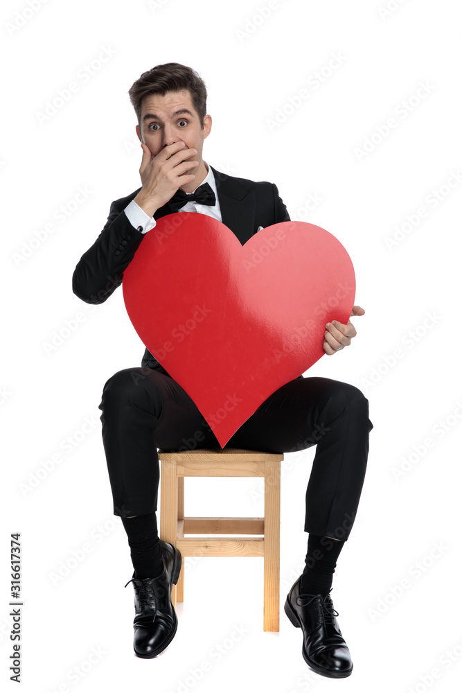 young elegant groom holding big red heart and covering mouth young elegant groom holding big red heart and covering mouth