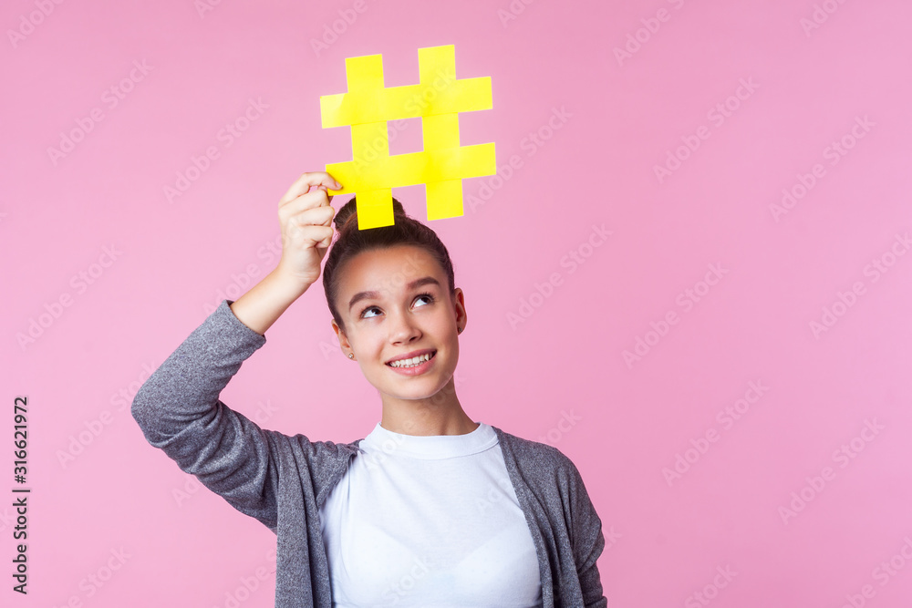 Blogging promotion. Portrait of trendy brunette teen girl with bun hairstyle in casual clothes smiling and looking at big hashtag symbol above her head. indoor studio shot isolated on pink background Blogging promotion. Portrait of trendy brunette teen girl with bun hairstyle in casual clothes smiling and looking at big hashtag symbol above her head. indoor studio shot isolated on pink background