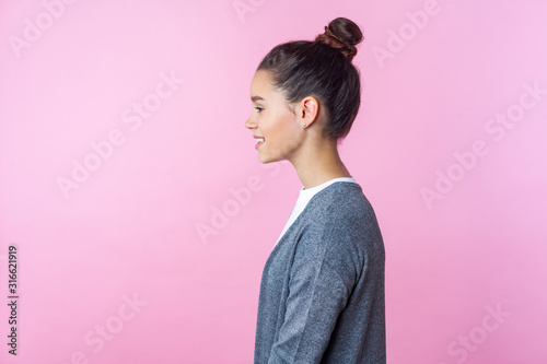 Side view of cheerful brunette teenage girl with bun hairstyle in casual pullover smiling, looking kind and positive, empty copy space for advertisement. indoor studio shot isolated on pink background
