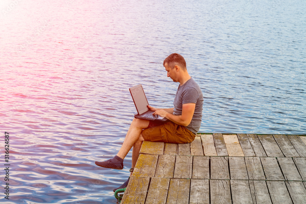 Young man sitting on a pier, using his laptop. Freelance job. Freedom lifestyle