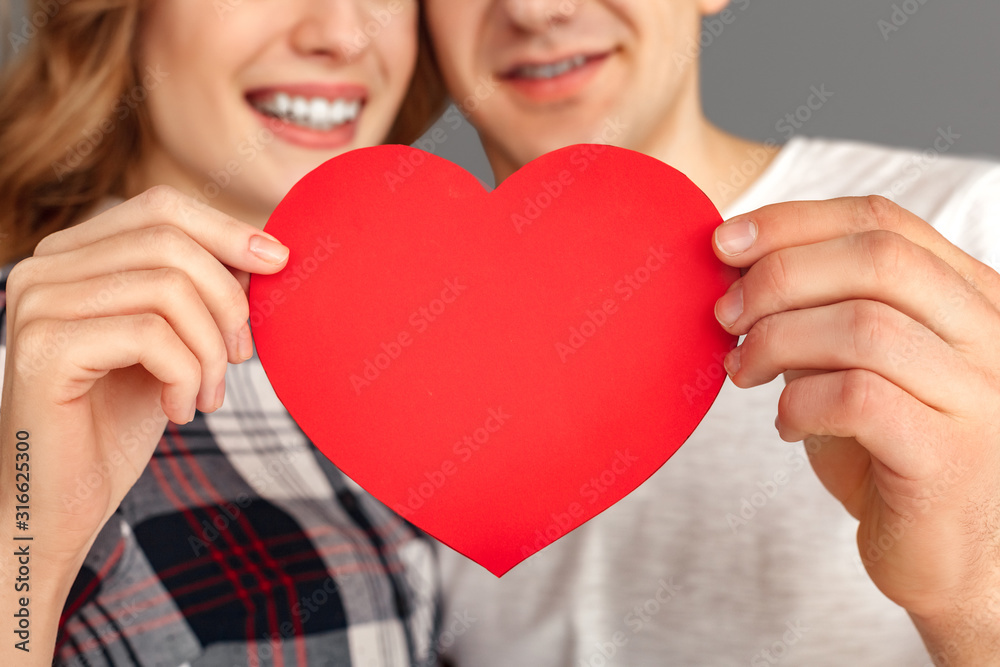 Valentines Day. Young couple standing isolated on grey with heart shape card close-up smiling happy blurred