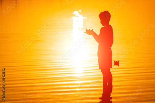 Little boy holding paper ship in hands. Floating ships into the distance. Beautiful sunset. Origami. River. Lake.