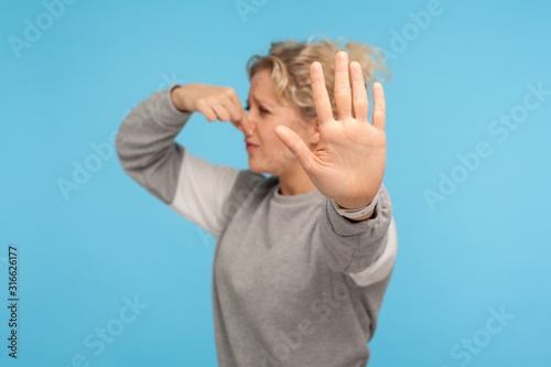 Bad breath. Woman in grey sweatshirt pinching nose with fingers, holding breath to avoid intolerable smell, stinky fart gases, showing stop gesture. indoor studio shot isolated on blue background