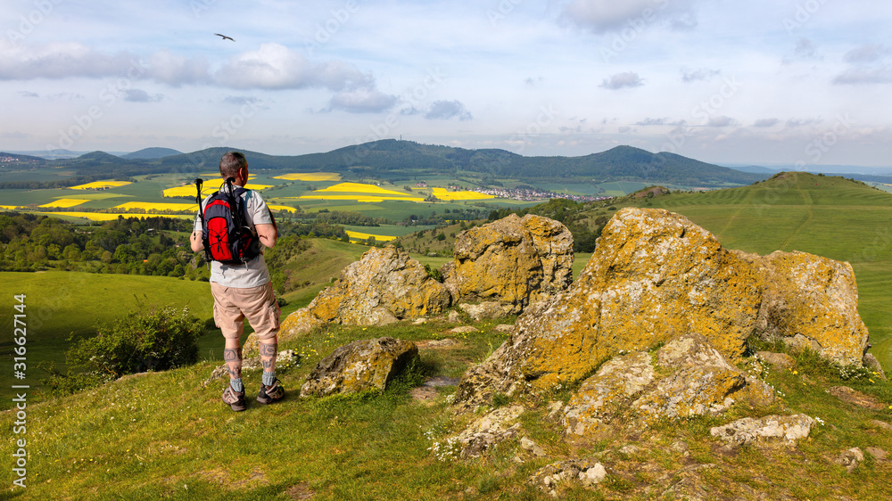 A hiker with a backpack near the city of Kassel on a mountain. The stones have the name Helfensteine. Below is an airfield for gliders. Rape fields bloom in the distance.