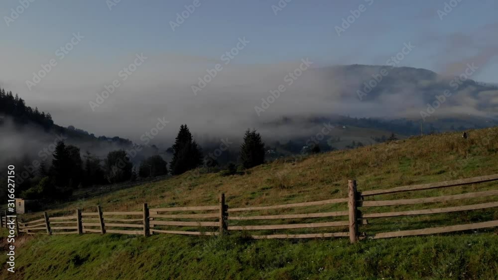Foggy morning in mountain village. Lath fence and village on green mountain slope. Scenic countryside scene. Foggy morning in mountain village. Lath fence and village on green mountain slope. Scenic countryside scene.
