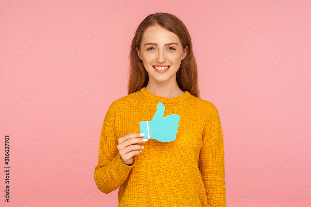 Like and share! Portrait of beautiful happy red hair girl in sweater holding blue thumb up love icon and smiling, social media recommendation, blogging. indoor studio shot isolated on pink background Like and share! Portrait of beautiful happy red hair girl in sweater holding blue thumb up love icon and smiling, social media recommendation, blogging. indoor studio shot isolated on pink background
