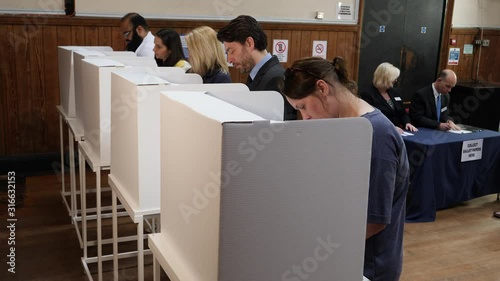 Voters voting at Polling Place / Station. People Stood in line at booths, choosing who to vote for. Ballot box behind.