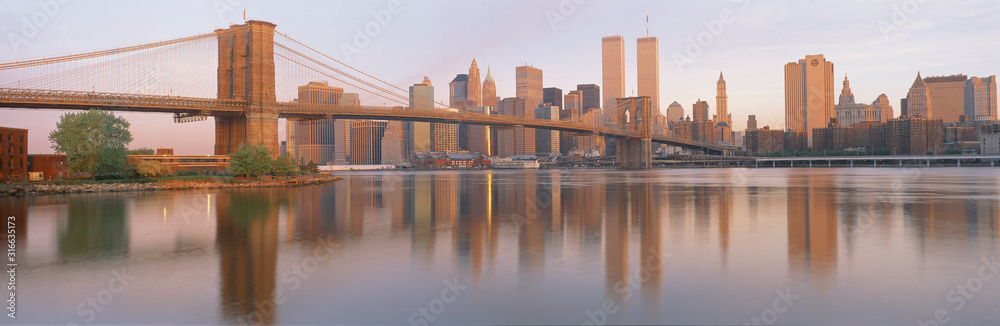 This is the Brooklyn Bridge over the East River. The Manhattan skyline is behind it at sunrise. The East River shows a reflection of the skyline.