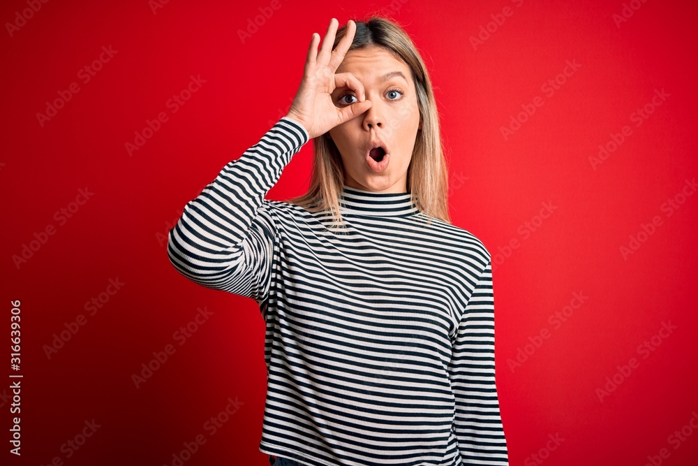 Young beautiful blonde woman wearing casual striped sweater over red isolated background doing ok gesture shocked with surprised face, eye looking through fingers. Unbelieving expression. Young beautiful blonde woman wearing casual striped sweater over red isolated background doing ok gesture shocked with surprised face, eye looking through fingers. Unbelieving expression.