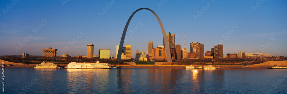 This is the skyline at sunrise. It is situated along the Mississippi River. There are riverboats on the water with the St. Louis Arch in clear view.