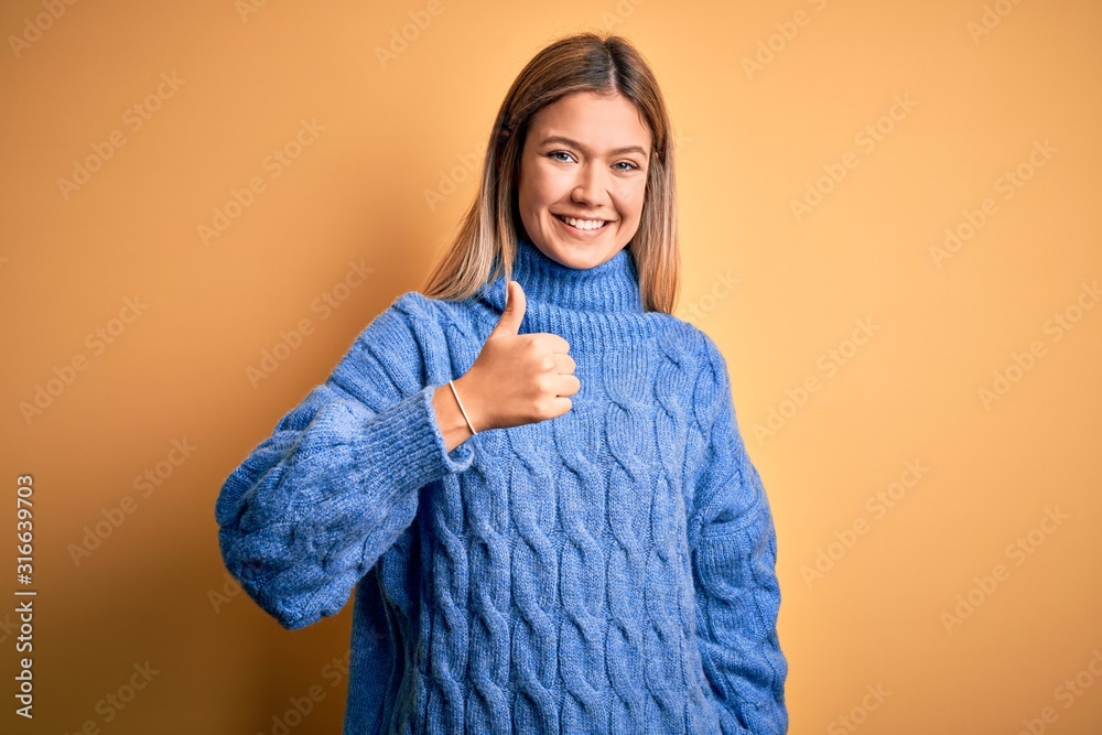 Young beautiful blonde woman wearing turtleneck sweater over yellow isolated background doing happy thumbs up gesture with hand. Approving expression looking at the camera showing success. Young beautiful blonde woman wearing turtleneck sweater over yellow isolated background doing happy thumbs up gesture with hand. Approving expression looking at the camera showing success.