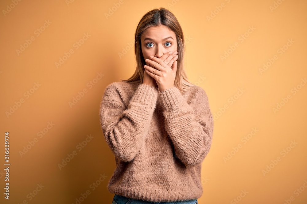 Young beautiful blonde woman wearing brown sweater over yellow isolated background shocked covering mouth with hands for mistake. Secret concept. Young beautiful blonde woman wearing brown sweater over yellow isolated background shocked covering mouth with hands for mistake. Secret concept.