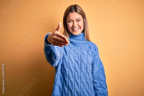 Young beautiful blonde woman wearing turtleneck sweater over yellow isolated background smiling friendly offering handshake as greeting and welcoming. Successful business.