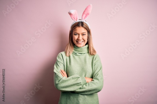 Young beautiful woman wearing easter rabbit ears standing over isolated pink background happy face smiling with crossed arms looking at the camera. Positive person.