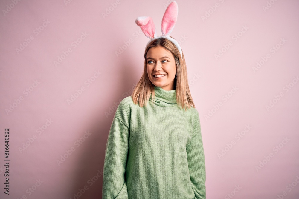 Young beautiful woman wearing easter rabbit ears standing over isolated pink background looking away to side with smile on face, natural expression. Laughing confident.