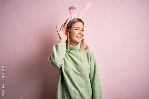 Young beautiful woman wearing easter rabbit ears standing over isolated pink background smiling with hand over ear listening an hearing to rumor or gossip. Deafness concept.
