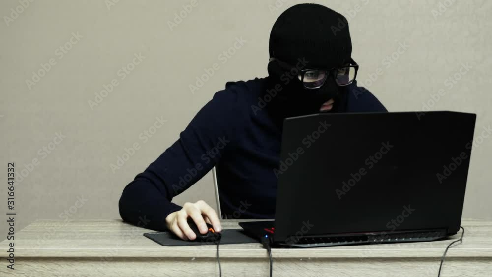 Funny male hacker in black mask balaclava puts on glasses and do hacker attack on laptop Funny male hacker in black mask balaclava puts on glasses and do hacker attack on laptop