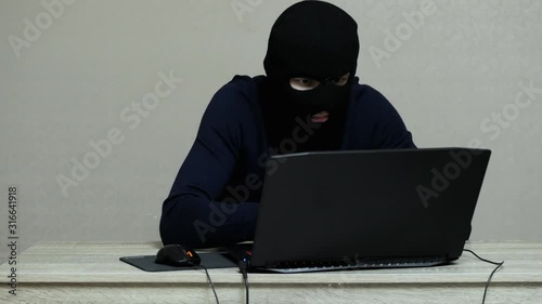Funny male hacker in black mask balaclava do hacker attack on laptop, looking at the camera and thumbs up