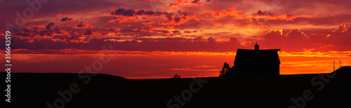 This is a farm at sunset. The farm house is silhouetted against an orange sky.