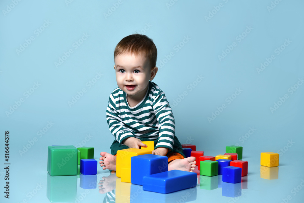 Little baby boy in stylish casual clothing barefoot sitting on floor and playing with colorful toy cubes over blue wall background Little baby boy in stylish casual clothing barefoot sitting on floor and playing with colorful toy cubes over blue wall background
