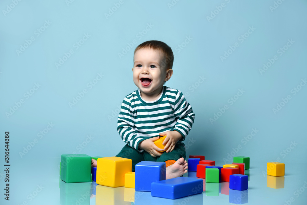 Little baby boy in stylish casual clothing barefoot sitting on floor and playing with colorful toy cubes over blue wall background Little baby boy in stylish casual clothing barefoot sitting on floor and playing with colorful toy cubes over blue wall background