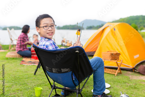 asian children holding grilled organic corn with hand, he camping in lakeside with his family, he sitting on seat and feeling happy, they relax in holiday, happiness family activity