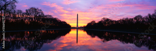 This is the Washington Monument at sunrise. The sky is a pink and purple with the monument reflected in its reflecting pool . It is surrounded by spring foliage.
