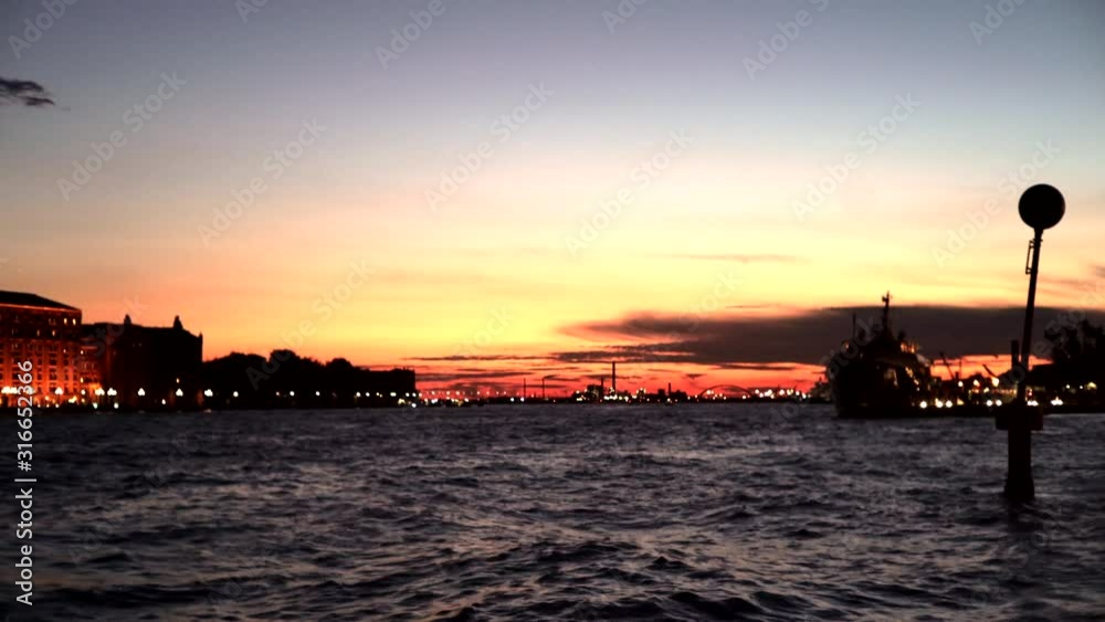 Gentle waves in Venice, wide shot of an orange sunset from the sea showing the cityscape with lights in the evening. Gentle waves in Venice, wide shot of an orange sunset from the sea showing the cityscape with lights in the evening.