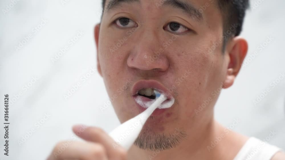 Back view of Asian guy in white shirt brushing teeth with an electric toothbrush in front of mirror in bathroom