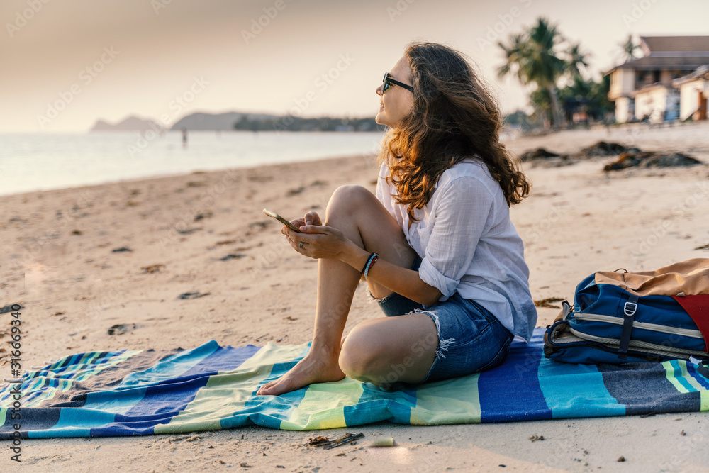 Young beautiful woman sits on the sand of a beach with a smartphone in her hands during sunset