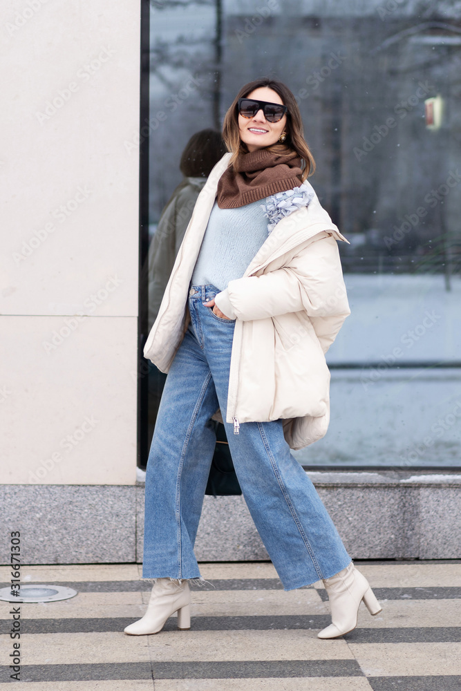 European model girl in a beige oversized down jacket, knitted sweater, flared jeans with a handbag and glasses is walking down the street. Life style