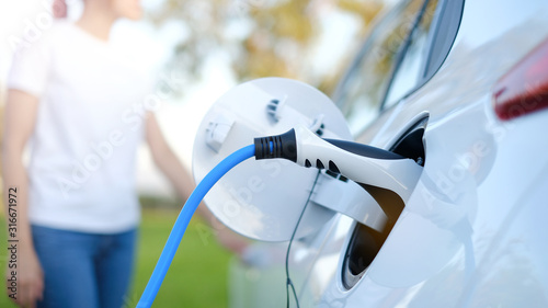 Electric car or ev is charging at station . man use the white power cable and plug on nature background. eco and clean energy concept with the mobile phone smart technology. zero gas no transmission