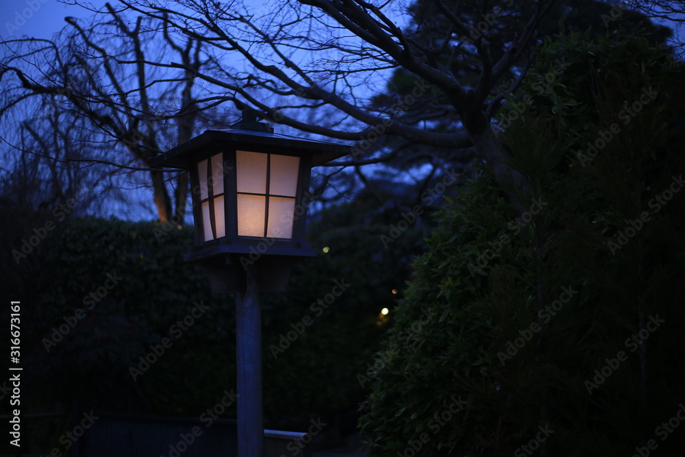 Japanese style garden lamp. Japanese gable roof house light post in traditional garden. warm light in cool sunset garden.