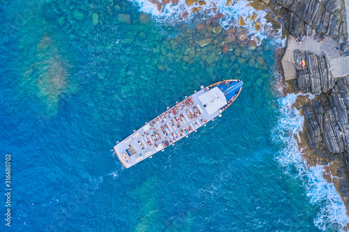 A white liner on blue water, the liner stopped at the rocky shore, aerial view, people on the deck. Cinque Terre National Park, Italy
