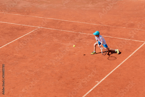 A boy plays tennis on an outdoor tennis court. The child is concentrated and focused on the game. Individual sport. Determined young athlete. Kids tournament, match. Active sport.
