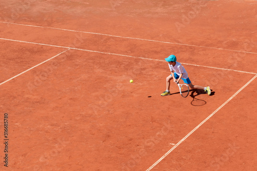 A boy plays tennis on an outdoor tennis court. The child is concentrated and focused on the game. Individual sport. Determined young athlete. Kids tournament, match. Active sport.
