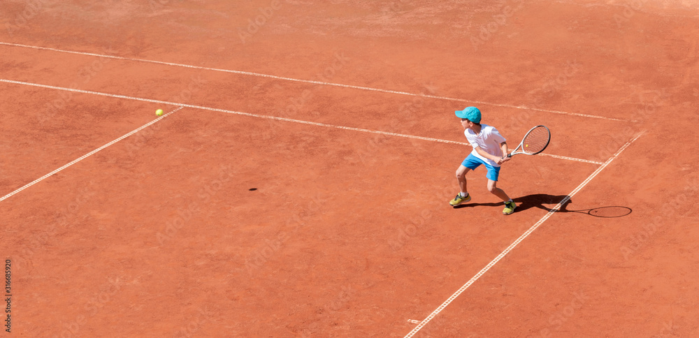 A boy plays tennis on a clay tennis court. The child is concentrated and focused on the game. Individual sport. Determined young athlete. Kids tournament, match. Active sport.