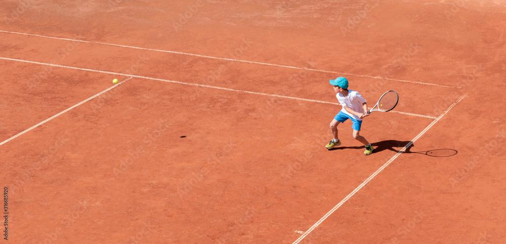 A boy plays tennis on a clay tennis court. The child is concentrated and focused on the game. Individual sport. Determined young athlete. Kids tournament, match. Active sport. A boy plays tennis on a clay tennis court. The child is concentrated and focused on the game. Individual sport. Determined young athlete. Kids tournament, match. Active sport.