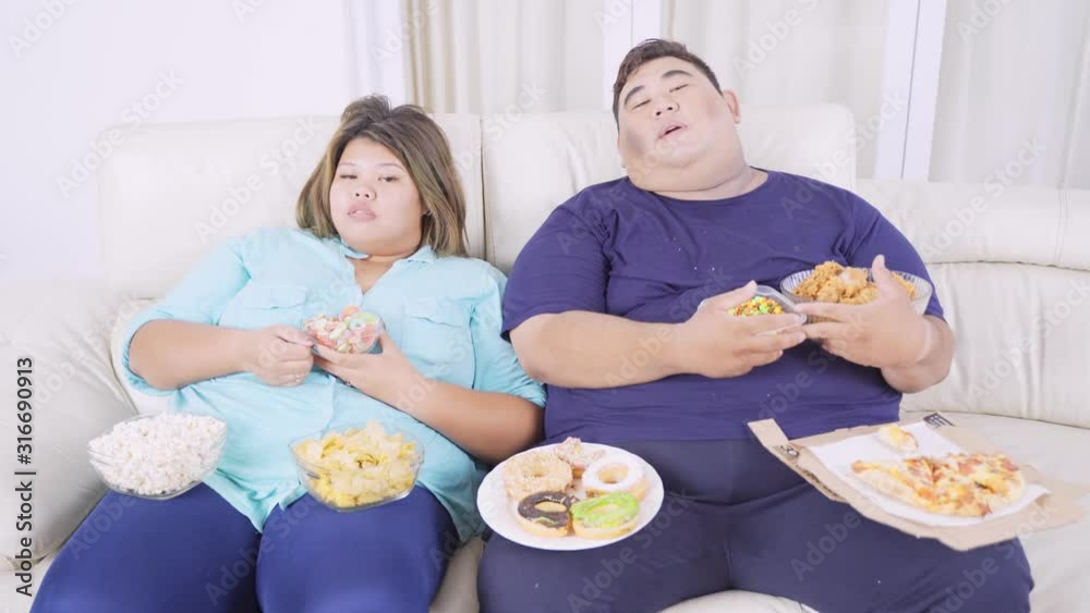 Obesity couple watching TV and looks asleep while eating junk foods in the living room at home. Shot in 4k resolution Obesity couple watching TV and looks asleep while eating junk foods in the living room at home. Shot in 4k resolution