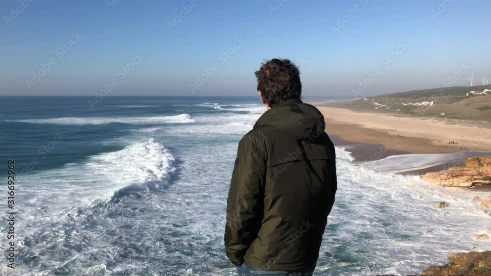 Young brunette man standing on steep cliff of Nazare coastline, enjoying a view of amazing crushing waves with white foam, drops of seawater; windy sunny evening; self-guided travel concept, Portugal Young brunette man standing on steep cliff of Nazare coastline, enjoying a view of amazing crushing waves with white foam, drops of seawater; windy sunny evening; self-guided travel concept, Portugal